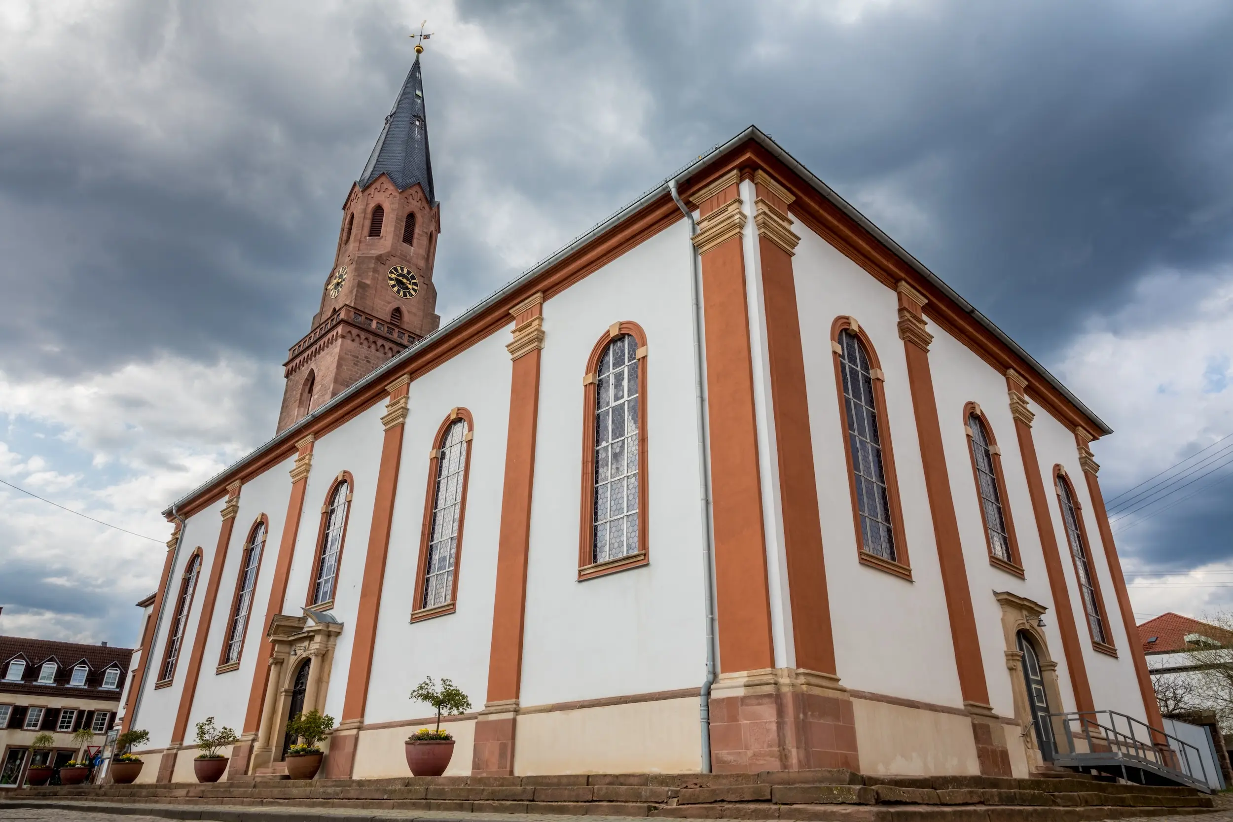 Blick auf das Kirchengebäude von Osten. Weiße Fassade mit braunfarbenen Säulen, dahinter der Kirchturm