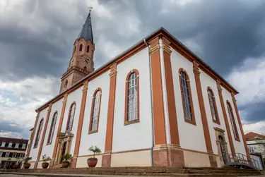 Blick auf das Kirchengebäude von Osten. Weiße Fassade mit braunfarbenen Säulen, dahinter der Kirchturm