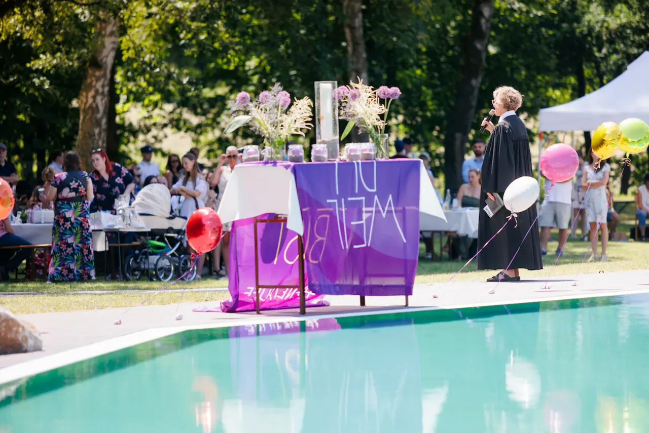 Tauffest im Naturschwimmbad Höhr-Grenzhausen: Eine Fahne der Taufinitiative der EKD liegt auf einem mit Blumen und Taufkerzen dekorierten Tisch. Daneben steht eine Pfarrerin mit einem Mikfrofon in der Hand - dahinter die Besucher*innen der Taufveranstaltung.