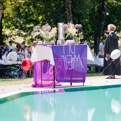 Tauffest im Naturschwimmbad Höhr-Grenzhausen: Eine Fahne der Taufinitiative der EKD liegt auf einem mit Blumen und Taufkerzen dekorierten Tisch. Daneben steht eine Pfarrerin mit einem Mikfrofon in der Hand - dahinter die Besucher*innen der Taufveranstaltung.