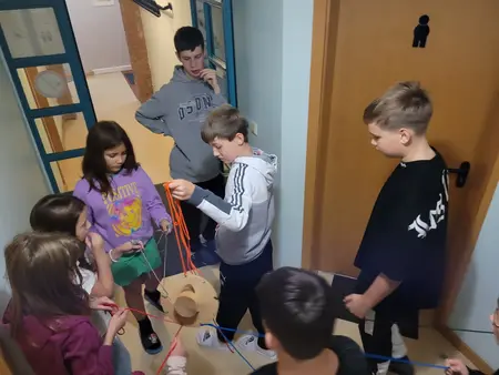 Kinder stehen im Flur, gemeinsam müssen sie mit Schnüren einen Gegenstand balancieren